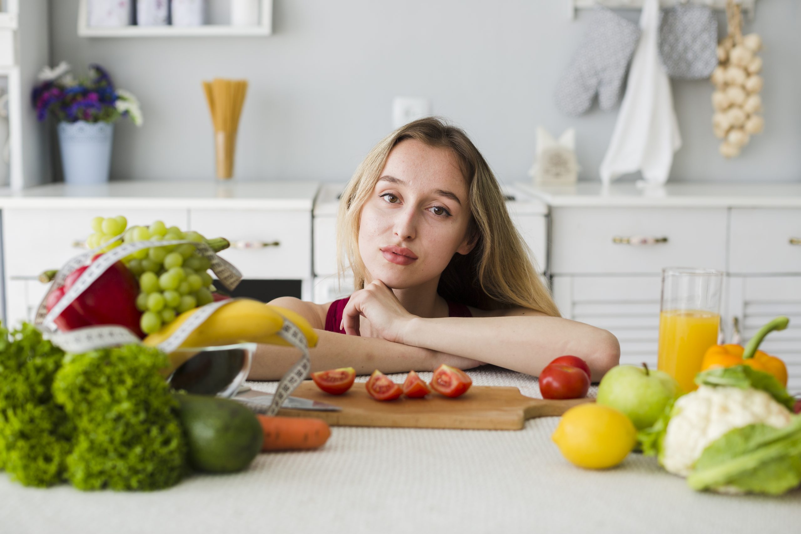 A brunette woman sits in the kitchen, seemingly trying to set her mindset, with fresh fruits and vegetables nearby.
