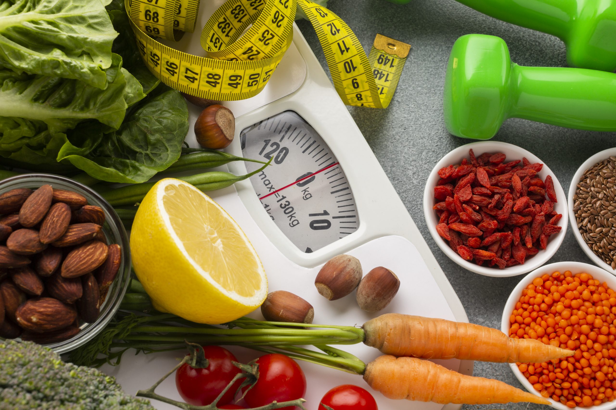 Healthy weight loss concept with weighing scale, measuring tape, vegetables, nuts, seeds, and dumbbell on table.
